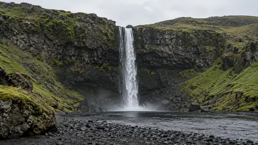 A tall narrow waterfall drops in one unbroken white ribbon over a dark cliff ledge into a black pebble shore pool, green moss on the cliff sides.