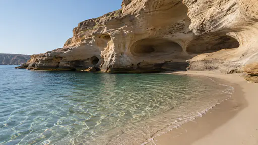 A wind-cut sandstone bluff with smooth scooped hollows rises above a small curved cove of pale sand and shallow water in bright side light.