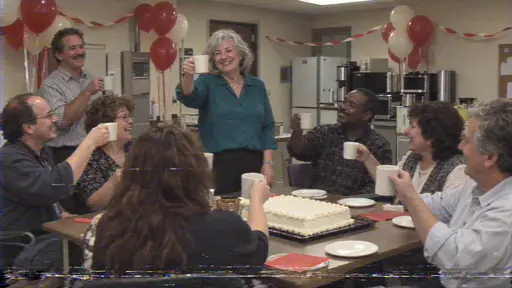 A fictional adult woman with silver hair in a teal blouse raises a mug of tea toward about eight adult colleagues around a break room table with a blank iced sheet cake and balloons in a camcorder still.