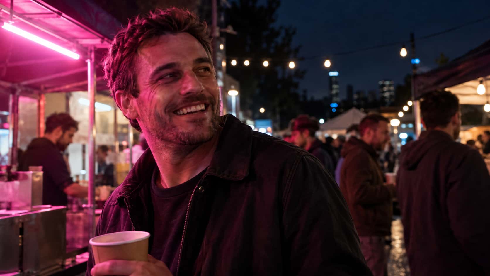 A fictional adult man with short hair and stubble in a dark jacket laughs warmly with a paper cup at an evening street food fair under a plain magenta neon tube.