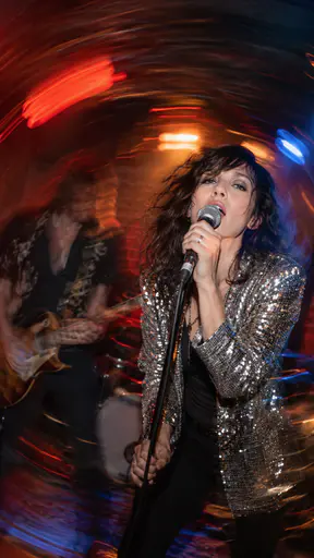 A fictional adult singer in a sequined jacket grips a microphone on a small club stage frozen sharp by a flash while the stage lights and a guitarist swirl into curved light trails.