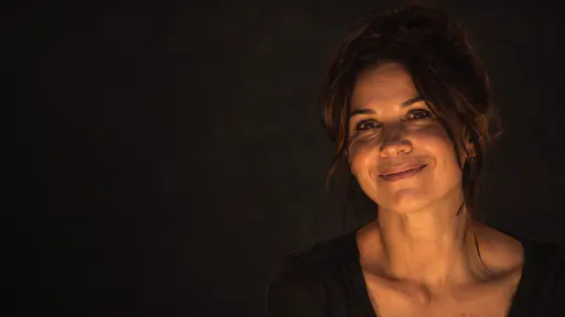 A fictional adult woman with a warm smile is lit from low in front below the eyeline against a deep charcoal background in a low key portrait photograph with fine grain.