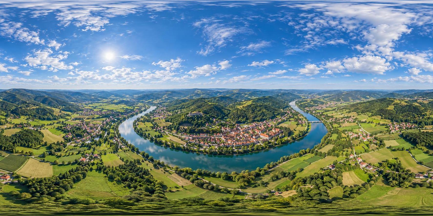 A full 360 degree equirectangular aerial panorama from high above a green river valley town with red roofed houses, a winding river, patchwork fields and forested hills with sky overhead.