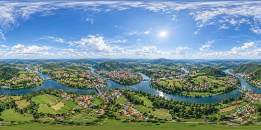 A full 360 degree equirectangular aerial panorama from high above a green river valley town with red roofed houses, a winding river, patchwork fields and forested hills with sky overhead.