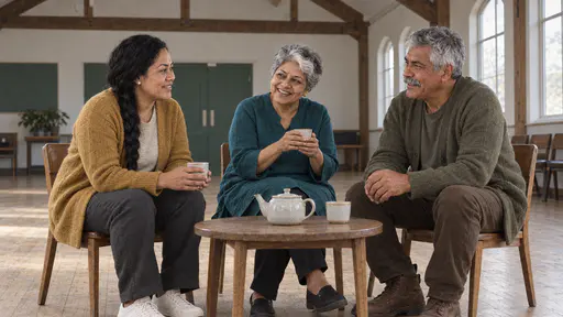 Three people, a woman with a long black braid, a silver-haired woman and a silver-haired moustached man, sit in a ring of wooden chairs around a small table with a teapot and three cups in a timber-beamed hall in diffuse afternoon light.