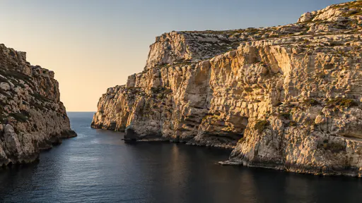 A bare arid cliff of layered pale rock drops straight to a narrow sea inlet of dark still water in warm afternoon light.