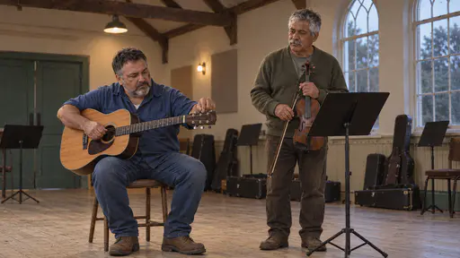 A stocky grey-bearded man in a blue shirt sits tuning an acoustic guitar while a silver-haired moustached man in an olive cardigan stands holding a fiddle beside a music stand with instrument cases along the wall of a timber-beamed hall in warm lamp light.