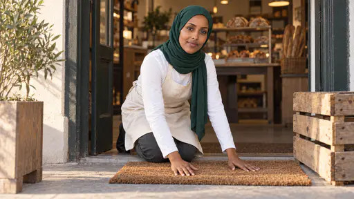 A woman in an emerald-green hijab and a cream apron kneels at the open front door of a bakery smoothing a coir welcome mat flat on the threshold with both hands, a wooden crate and a potted olive tree beside the door and loaves on shelves inside.