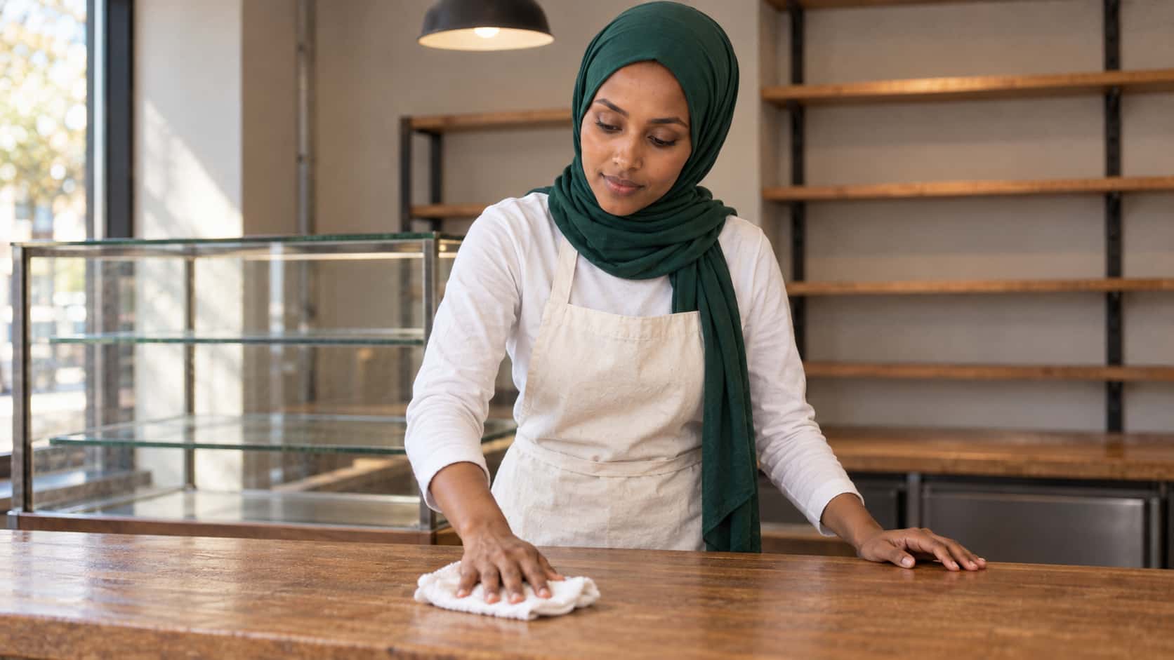 A woman in an emerald-green hijab, a cream apron and a white top wipes a bare wooden bakery counter with a white cloth, an empty glass display cabinet, bare shelves and an overhead lamp behind her.