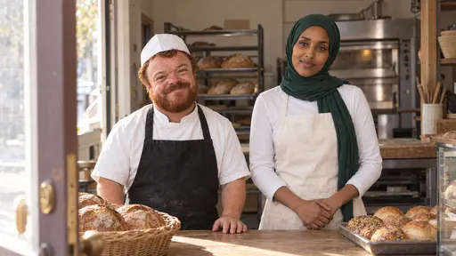 A baker with a thick red-brown beard and a white paper cap, in a white jacket and black apron, and a woman in an emerald-green hijab and a cream apron stand behind a wooden counter smiling, a basket of loaves and a tray of seeded rolls on the counter and morning light coming through an open door.