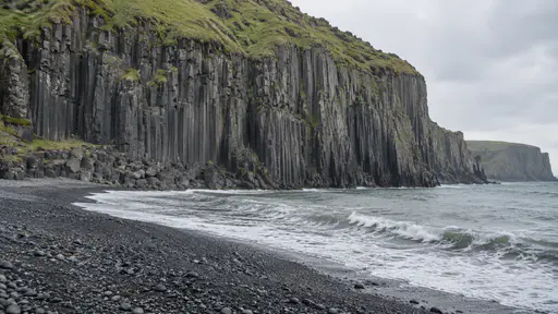 A wall of tall straight black basalt columns rises from a black pebble shore into a grassy cliff top in bright side light, grey sea at its foot.