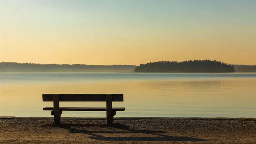 Solitary wooden bench beside a calm lake captured as a 16:9 landscape photograph with soft golden-hour light and clean composition for wallpaper use.