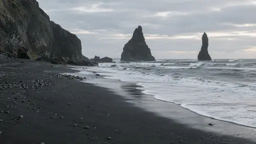 A black sand beach stretches along a grey sea with white foam lines in pale morning light, dark sea stacks standing offshore.