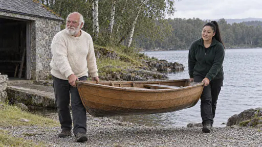 A broad white-bearded man in a cream jumper and a broad-shouldered woman with a high black ponytail in a green fleece carry a small wooden rowing boat down a gravel slipway toward a calm lake and birch trees in bright morning light.