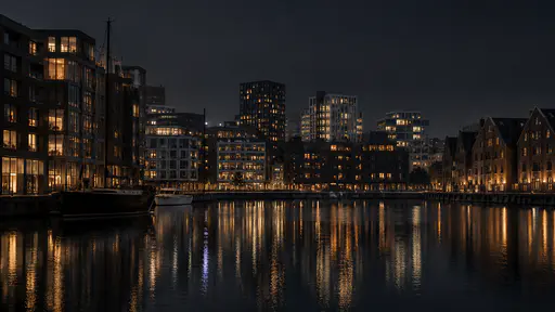 Clustered dark buildings with lit windows gather around a still harbour inlet at night, their broken reflections held in the black water.