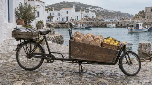 A cargo bicycle with a long wooden front box packed with bread in paper bags and a basket of lemons and a crate of fish on the rear rack stands on its kickstand on a cobbled harbour lane in early morning with whitewashed houses behind.