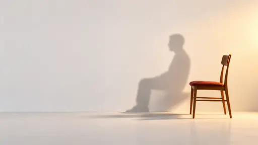 Minimal conceptual Halloween background: a single wooden chair with a muted-red cushion stands in right-of-center side profile against a seamless chalk-white studio wall, lit by soft amber light from the right. Beside it falls the small softly blurred charcoal shadow of a seated guest — rounded head, sloped shoulders, bent knees — though no one is there. The left two thirds of the wall stay undisturbed as clean copy space.