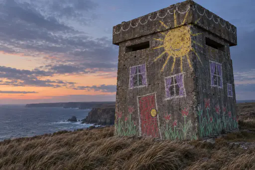 A squat gray concrete watchtower on a grassy sea cliff at dusk, covered in oversized crude chalk drawings: a wobbly yellow sun, crooked windows, a leaning red door and green flower scribbles.