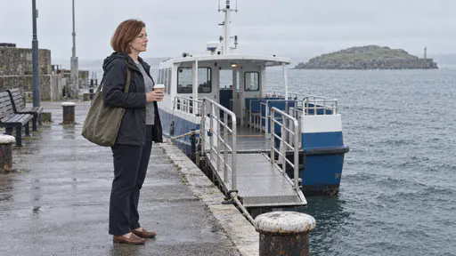 A woman with a chin-length auburn bob and a canvas work bag on her shoulder holds a paper coffee cup at the head of the gangway of a small blue and white ferry on a mainland quay in cool early light.