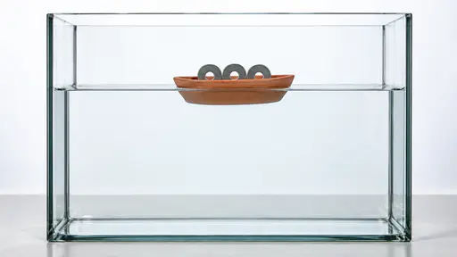 A hollow clay boat floats upright at the waterline carrying three steel washers and riding lower in a tall clear glass tank of water in front of a bright diffused backdrop.