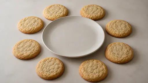 Several plain uniced sugar cookies are arranged in a loose ring around one empty plain plate on a plain table in even daylight.