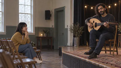 A slim bearded musician plays an oud on a low stage with his eyes closed while a petite young woman with a dark fringe in a mustard cardigan sits in the front row with her hands clasped under her chin and a rapt smile.