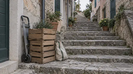 Three stacked plain wooden shopping crates, a folded metal handcart and a canvas shopping bag are parked on a mid-flight stone stair landing between shuttered house fronts in soft diffuse morning light.