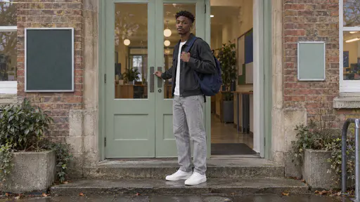 A tall young man with a coily twist-out in a charcoal jacket and navy rucksack hesitates on a stone step with one hand lifted toward the handle of a pale green double door in grey late-afternoon light.