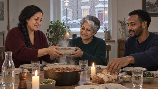 A Samoan woman passes a steaming bowl to a silver-haired woman while a tall bearded man reaches for a loaf of bread at a candle-lit dining table with a frosted window behind them.