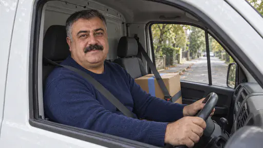 A heavyset man with a salt-and-pepper crew cut and wide moustache in a navy sweater drives a plain white van down a quiet tree-lined street with a sealed medium brown parcel held by a seat belt on the passenger seat seen through the open window in morning light.