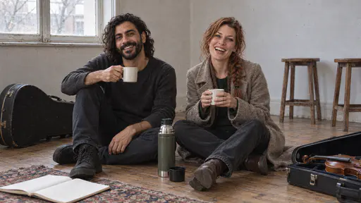 A slim bearded man and a freckled woman with a loose auburn braid sit on the wooden floor of a small rehearsal room with their backs to the wall, laughing and sharing tea from two mugs, a guitar and a violin resting on their cases beside them in low afternoon light.