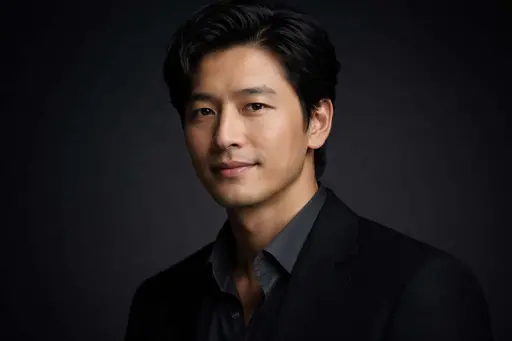 Wide 3:2 cinematic studio headshot of an East Asian man in his early 40s, collarbone-to-above-head crop, three-quarter turn with a warm restrained smile against a near-black gradient backdrop.