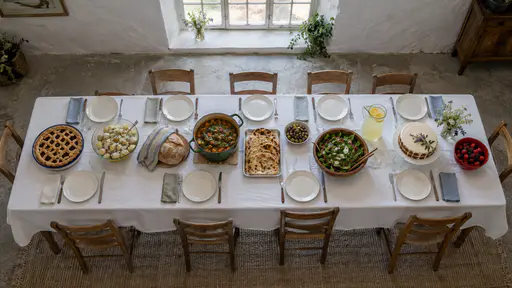 A long trestle table seen from above is laid with a lattice pie, a bowl of potato salad, a loaf, a green stew pot, a tray of flatbreads, olives, a bowl of salad, a lemonade jug, an iced cake and a bowl of berries.