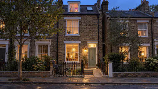 A narrow three-storey brick terrace glows from every floor at blue dusk with a lit ground-floor window beside a sage green door and stone ramp, a small coloured lamp in the attic window and a bicycle with a child seat chained to the railing.