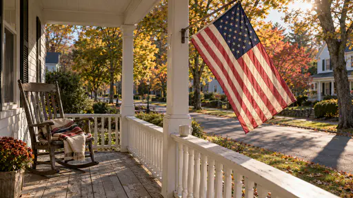 A faded American flag hangs from a bracket on a white clapboard porch post above an empty wooden rocking chair with a folded blanket and a cup on the rail in a slide film photograph.