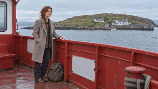 A freckled woman with a loose auburn braid in an oatmeal coat stands at the rail of a small red ferry with her rucksack at her feet and looks toward a small green treeless island with whitewashed cottages and a grey stone harbour in cool morning light.