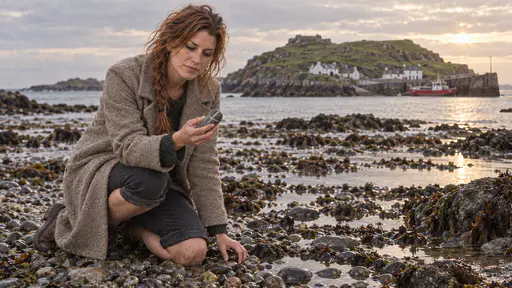 A freckled woman with an auburn braid kneels at the tideline of a pebble beach at low tide turning over a smooth grey striped stone beside rock pools with the small green island and whitewashed cottages behind in golden late-afternoon light.