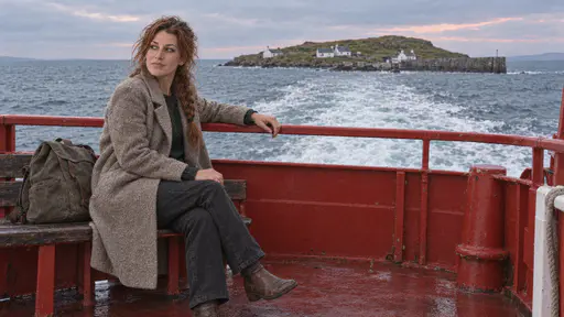 A freckled woman with an auburn braid sits on a bench on the stern deck of a small red ferry at dusk with her rucksack beside her and one hand on the rail looking back at the small green island with whitewashed cottages in the wake.