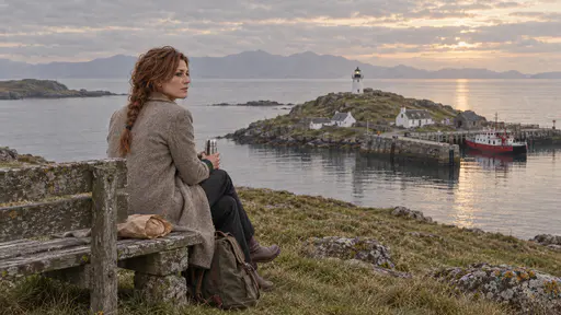 A freckled woman with an auburn braid sits on a weathered wooden bench on a grassy headland with a steel flask on her knee and a paper-wrapped loaf beside her looking across glassy water toward the mainland haze in warm evening light.