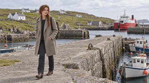 A freckled woman with an auburn braid and a rucksack walks slowly along a grey stone harbour wall past small moored fishing boats with whitewashed cottages on the grassy slope beyond in bright noon light.