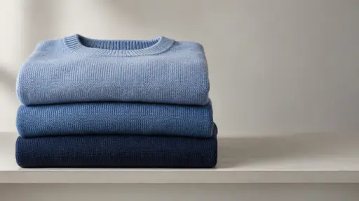 A neat stack of folded blue knitwear in three plain shades rests on a plain shelf edge in gentle window light.