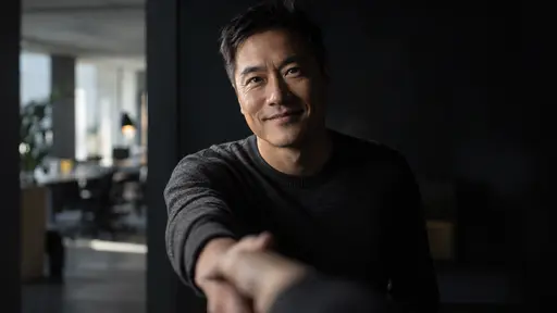 A fictional adult Chinese American founder, a man with short black hair in a dark grey sweater, extends a hand in welcome toward the camera with a warm smile against a deep charcoal field.