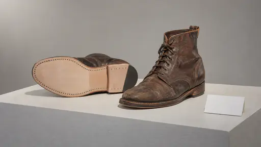 Worn leather boots on a museum plinth, one tipped on its side showing a pale new stitched replacement sole and heel, with a blank label card beside them.