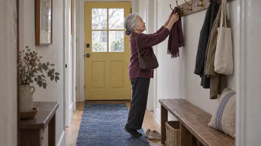 A very small silver-haired elderly woman with a low bun stands on tiptoe lifting a folded plum scarf off a brass coat hook in a narrow hallway with a shoulder bag on her arm.