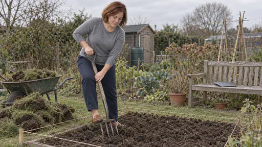 A woman with auburn bob in a heather-grey sweater turns over dark soil in a marked rectangle of lawn with a garden fork, a pile of cut turf beside the bed and a closed laptop on a garden bench nearby in overcast daylight.