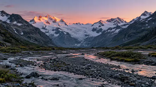 Braided glacial meltwater spreads across a gravel flat with low alpine willow and moss in the foreground while snowy ridgelines glow in rose and amber alpenglow in a mountain landscape photograph.
