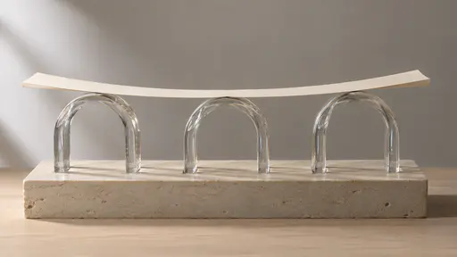Three clear glass arch segments stand side by side on a pale stone plinth like a short viaduct and carry a long curved cream paper roadway with a folded edge laid along their tops, each arch crown touching the underside of the road.