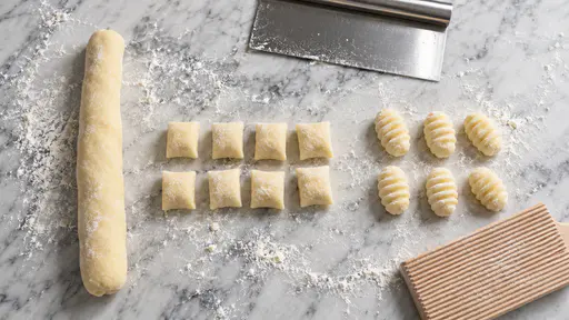 From above on floured marble, a potato dough rope at the left, eight cut pillows in two rows in the centre, six ridged gnocchi at the right, a beechwood ridged gnocchi board at the lower right and a steel dough scraper at the top.