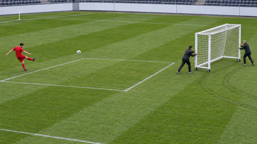Moving the goalposts: a soccer ball frozen mid-flight heading for an empty chalked goalmouth on green grass while two officials roll the whole white goal sideways on small wheels, leaving fresh curved tracks in the turf.