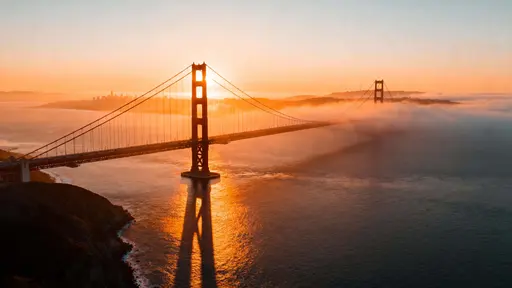 Aerial 16:9 composition of the Golden Gate Bridge at sunrise, orange sunlight piercing morning mist with long shadows and calm bay reflections in ultra-realistic warm tones.